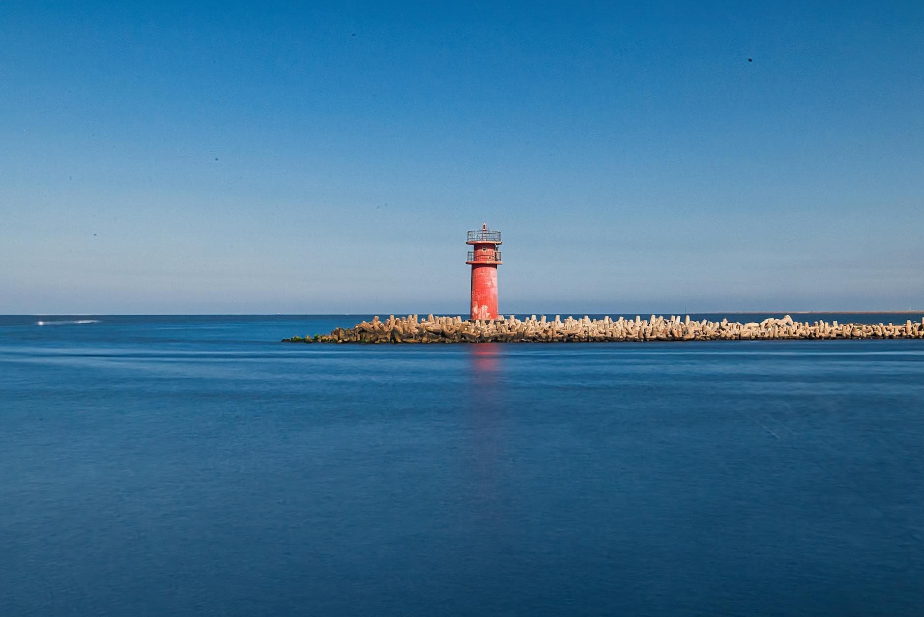 A vibrant red lighthouse stands on a tranquil coastline in Ras El-Bar, Egypt, under a clear blue sky.