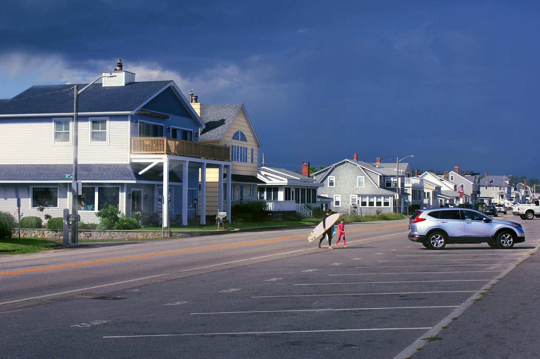 A charming coastal street view with houses, cars, and a surfer carrying a surfboard in New Hampshire.