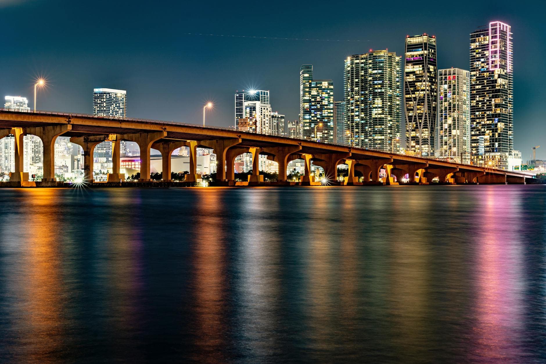 Illuminated Miami skyline with a bridge reflection at night.