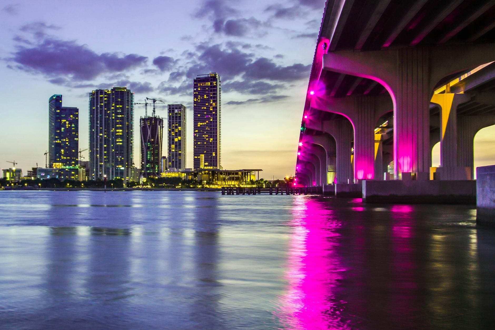 Captivating view of Miami skyline and illuminated bridge reflecting on water at dusk, vibrant city lights aglow.