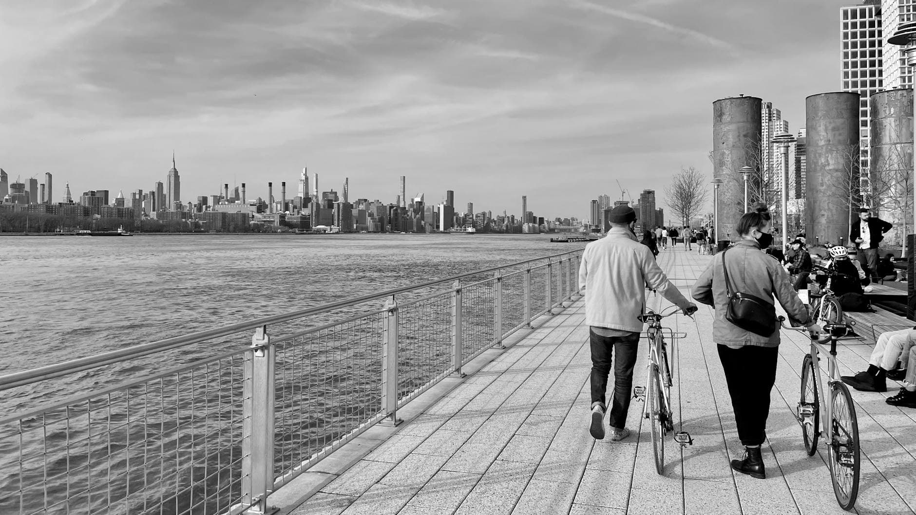 People walking with bicycles on a New York waterfront promenade overlooking the skyline.