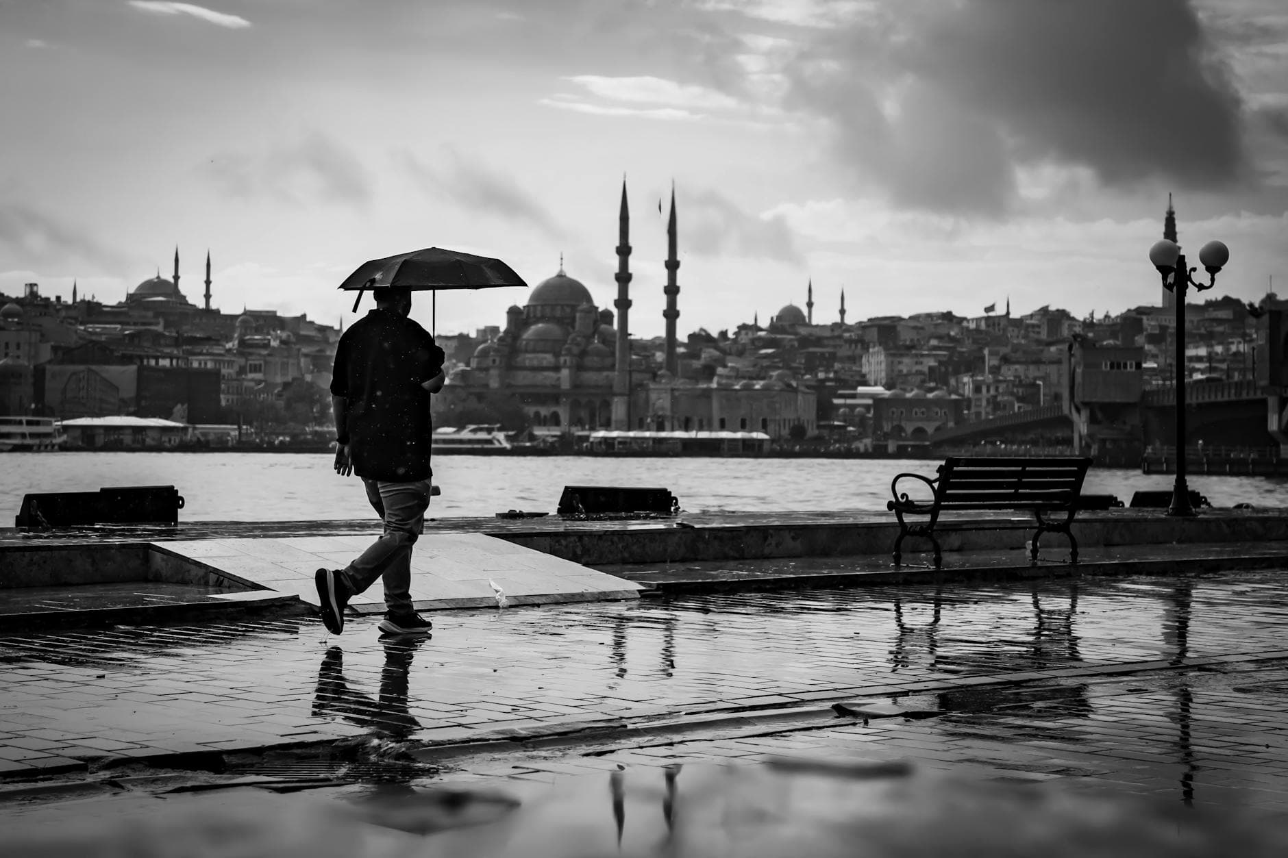 A person walks with an umbrella along İstanbul's waterfront, featuring a classic skyline view.
