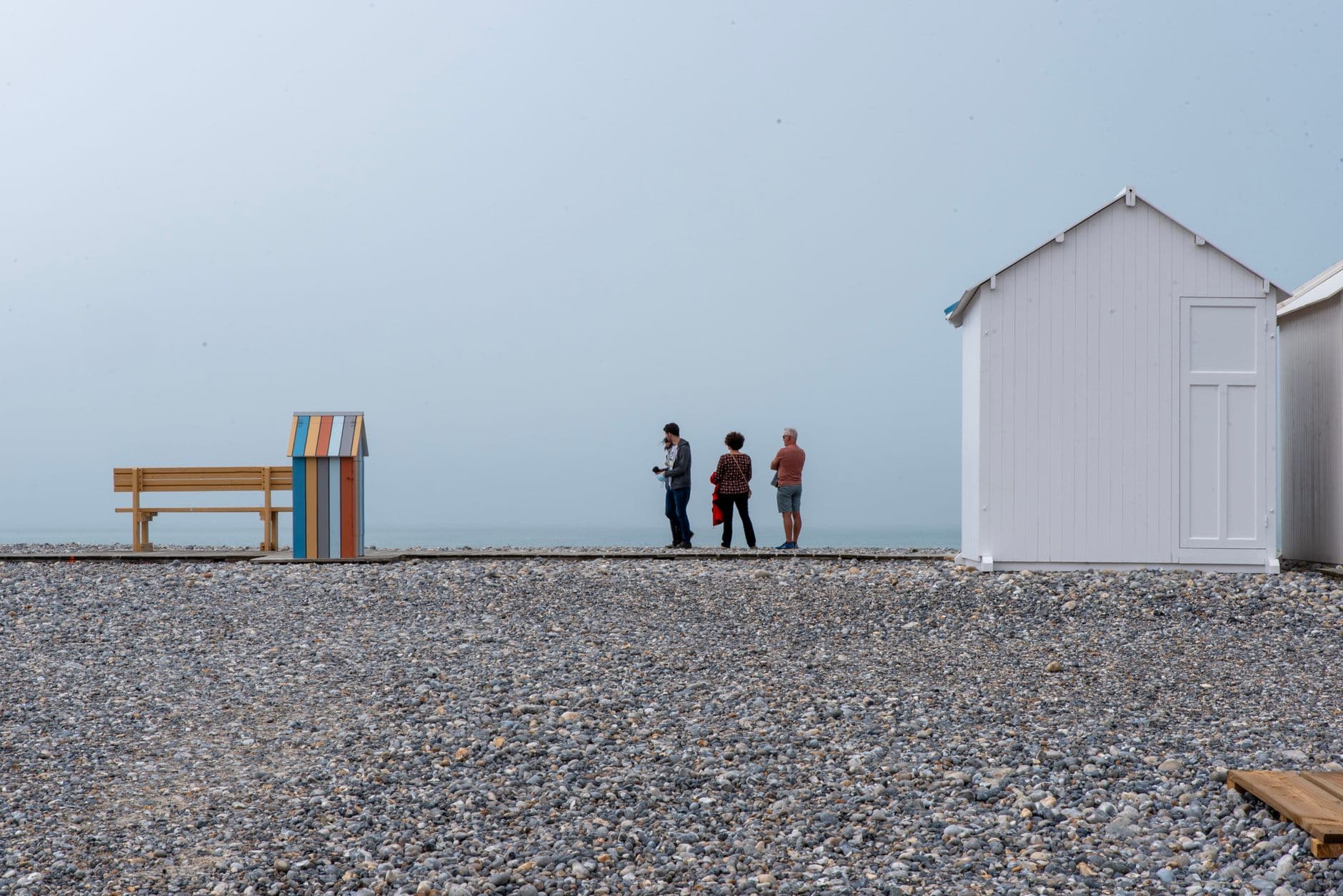 A serene pebble beach in Cayeux-sur-Mer, France, featuring colorful bench huts and people enjoying the view.