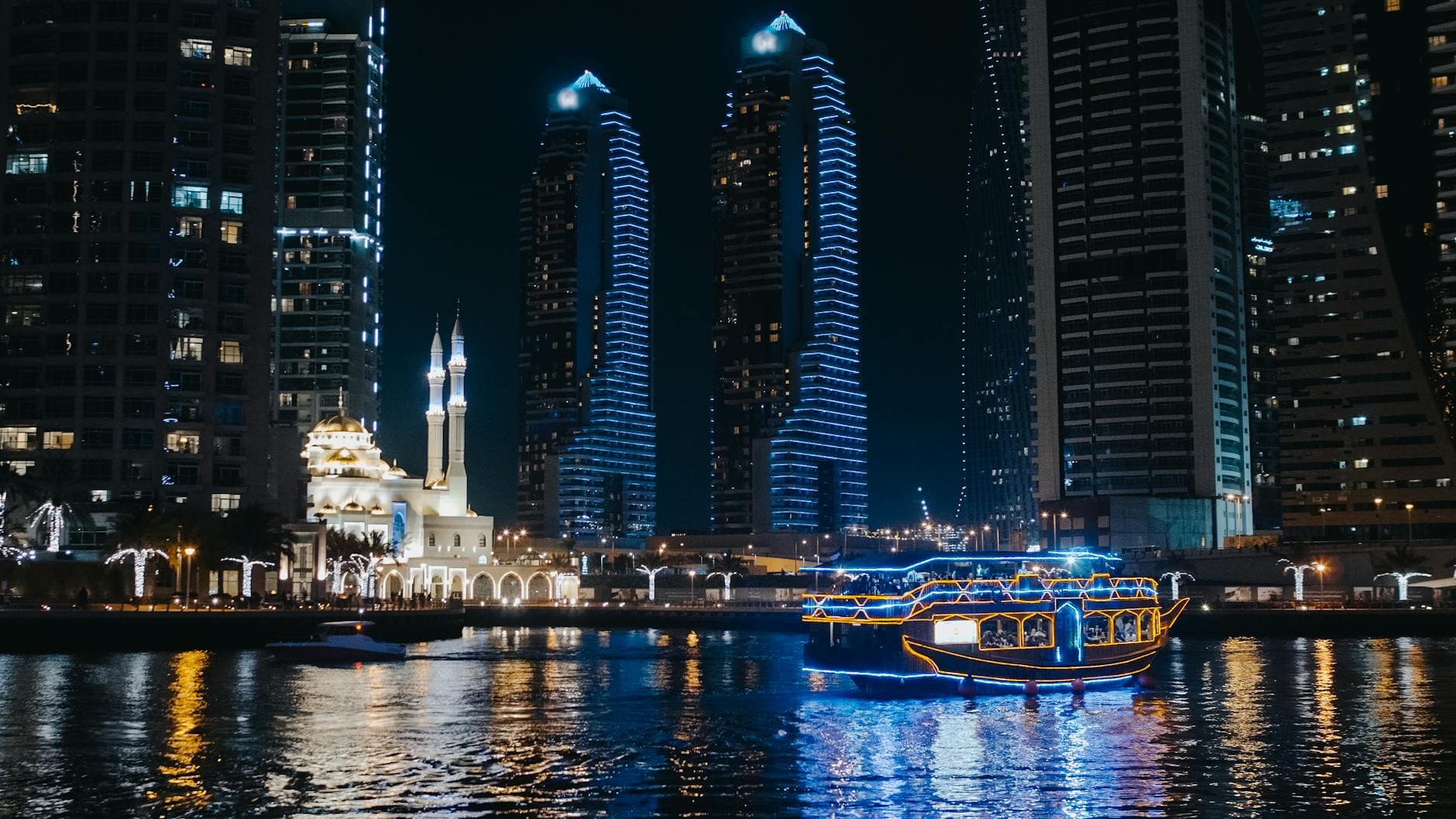 Illuminated skyscrapers and mosque reflecting in Dubai Marina with a vibrant boat.