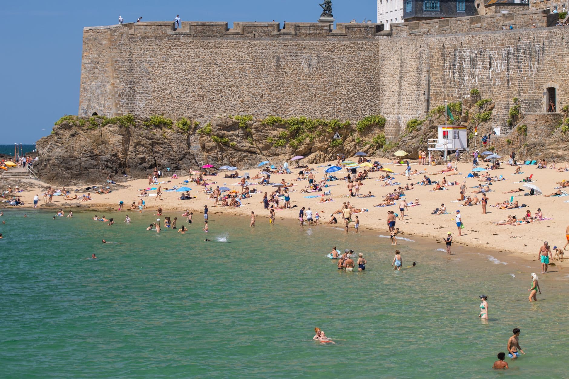 Tourists enjoy a sunny day on the beach in Saint-Malo, France, with historic fortifications in view.